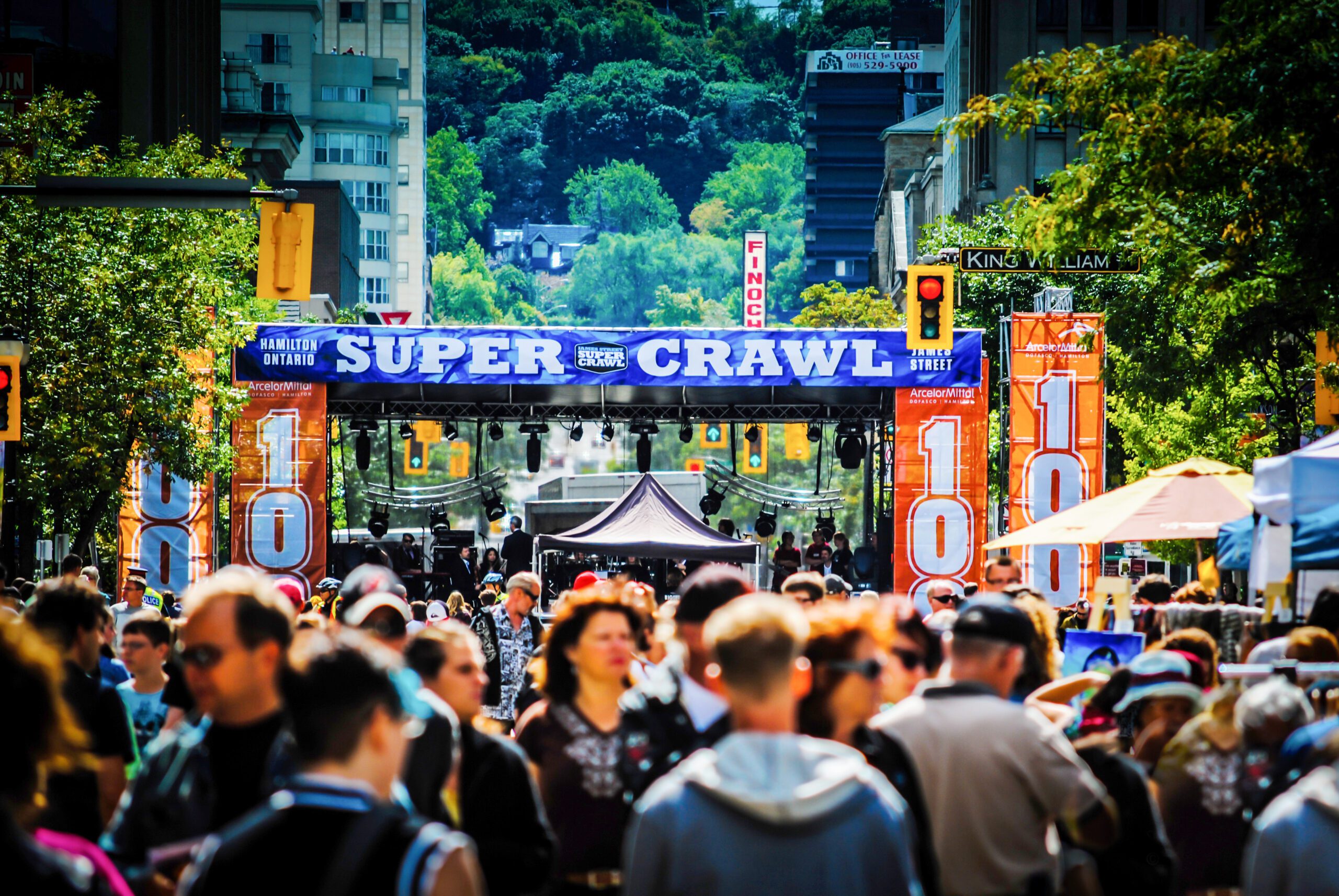A bustling street festival with a large "Super Crawl" banner, crowded with people and surrounded by trees and tall buildings.
