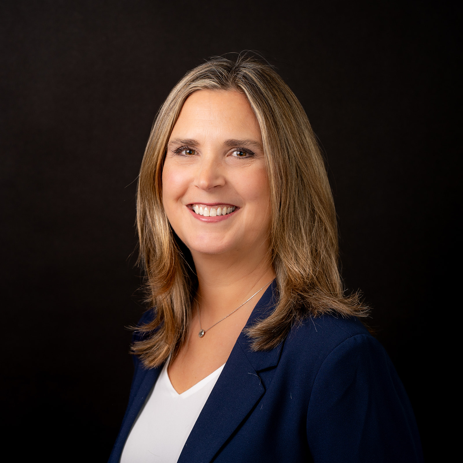 Woman with shoulder-length blonde hair in a blue blazer and white top smiles against a plain black background.
