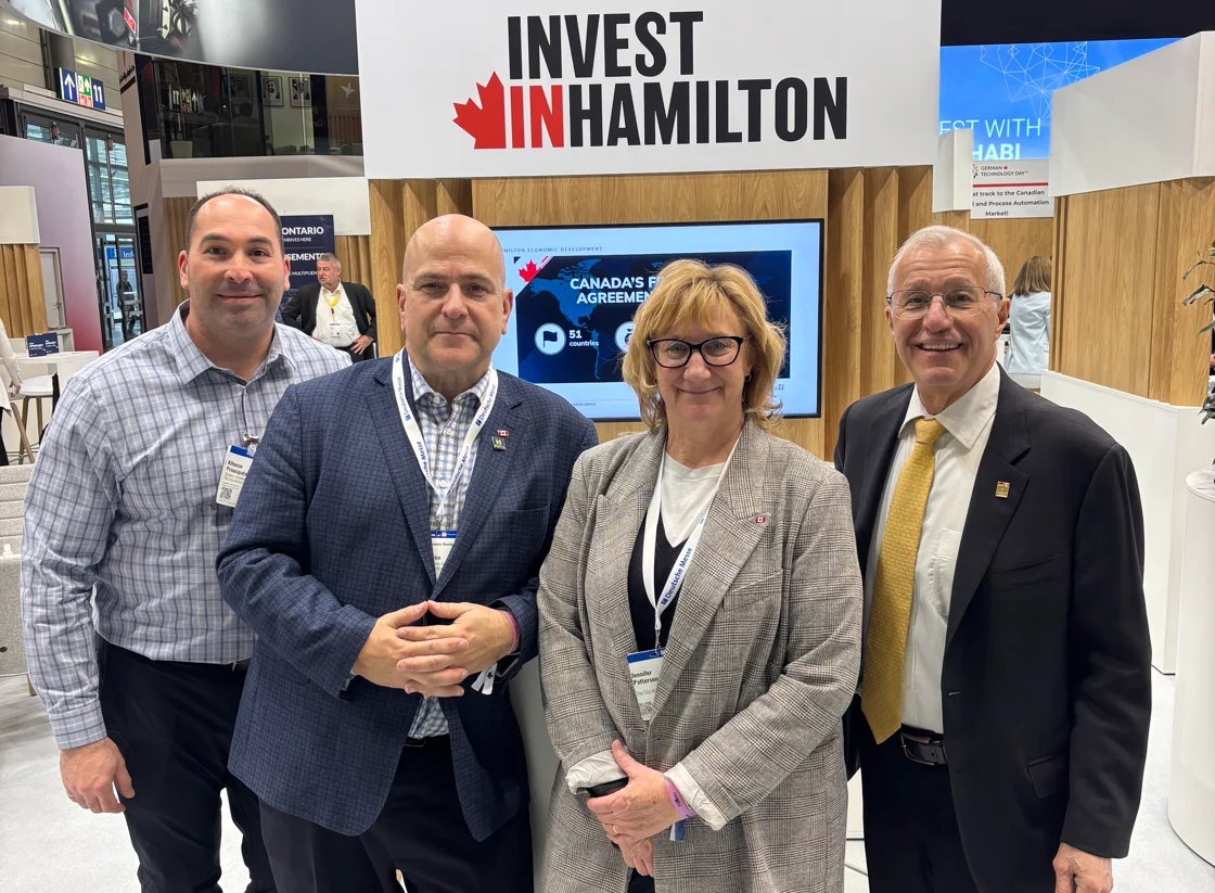 Four people stand in front of an "Invest in Hamilton" sign at a business event, facing the camera and smiling.