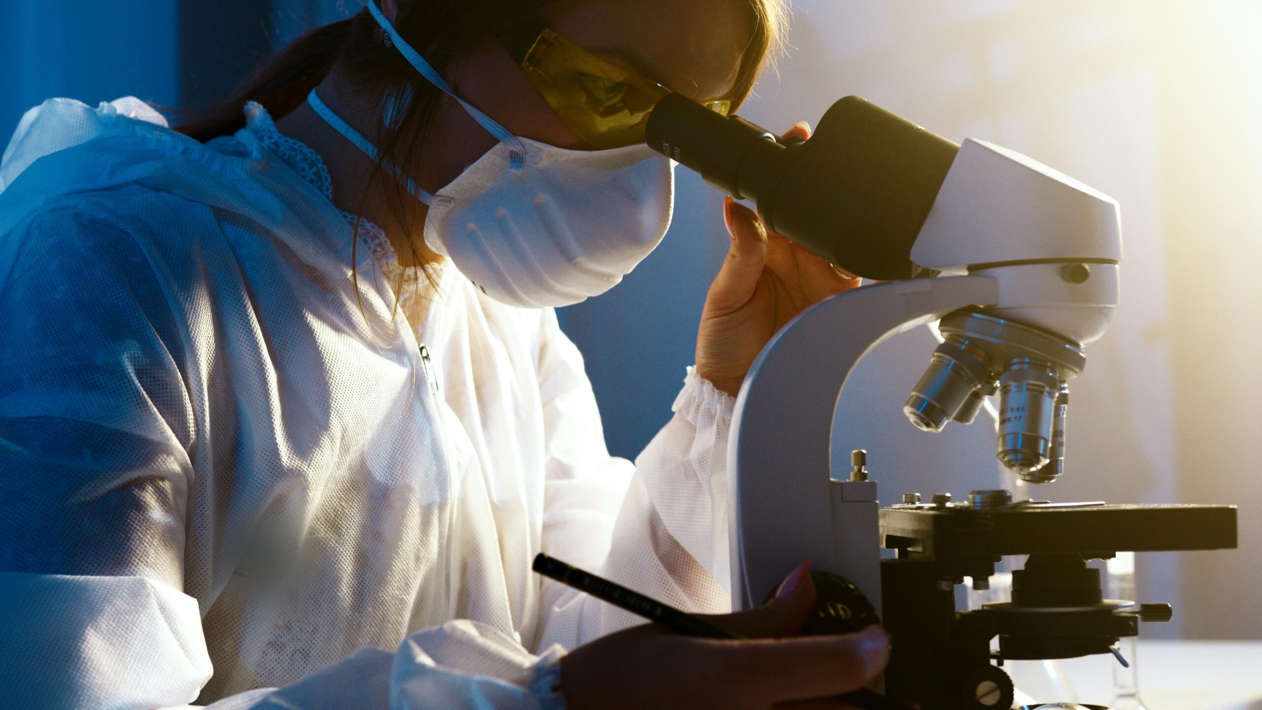 A person in protective clothing and a mask examines samples through a microscope in a laboratory setting.