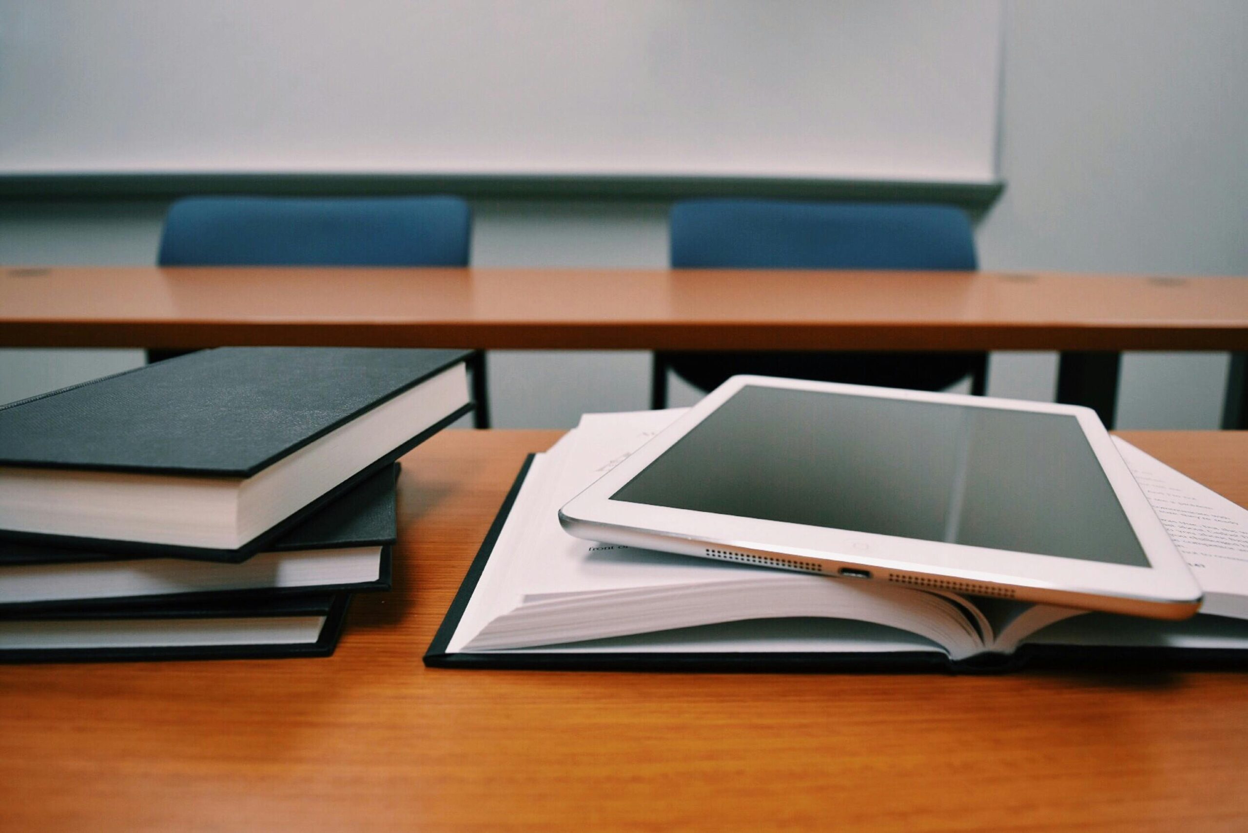 A tablet rests on an open notebook beside stacked books on a classroom desk with empty chairs in the background.