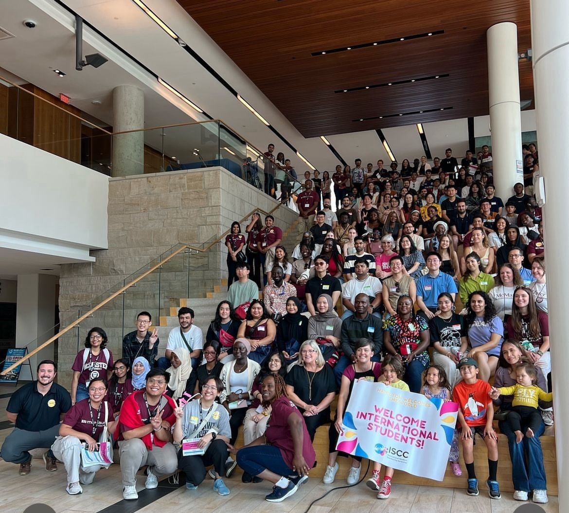 A large group of people poses on indoor stairs, holding a colorful "Welcome International Students" sign.