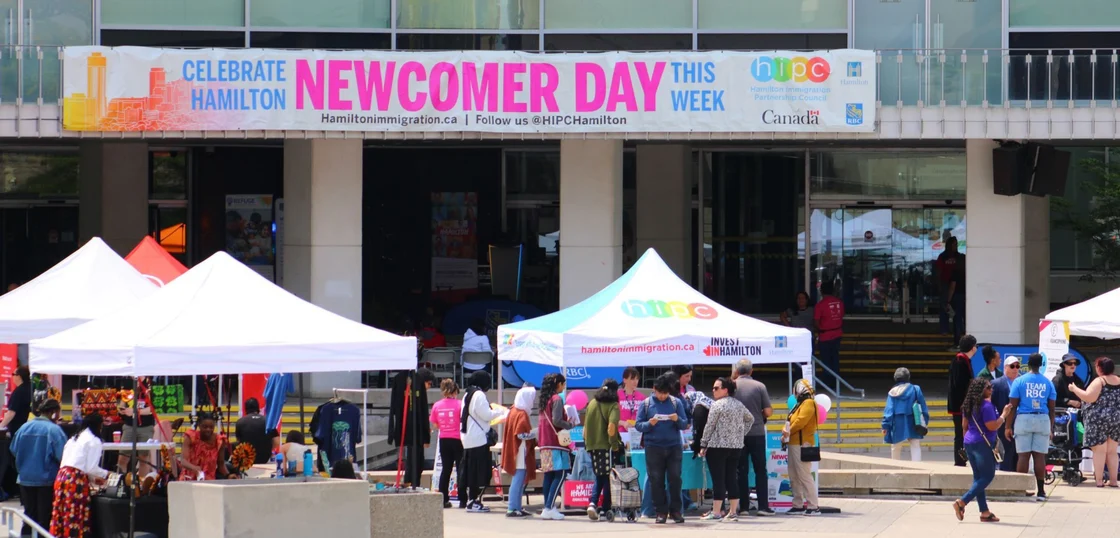 People gather outside a building with tents for Hamilton Newcomer Day, as seen on a banner overhead.