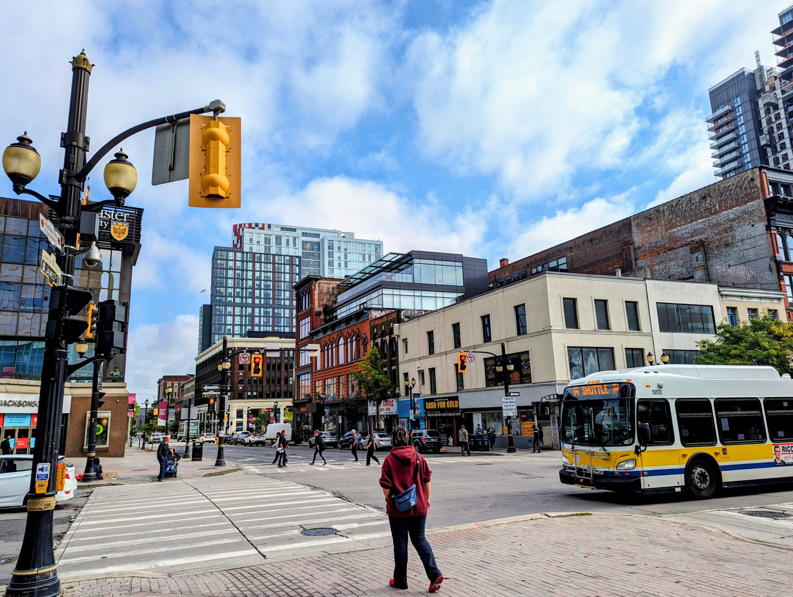 City intersection with pedestrians crossing, a bus stopped at the light, and modern buildings in the background.