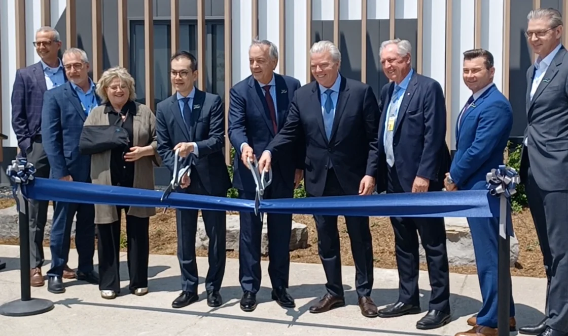 A group of people in business attire cut a blue ribbon at an outdoor ribbon-cutting ceremony.