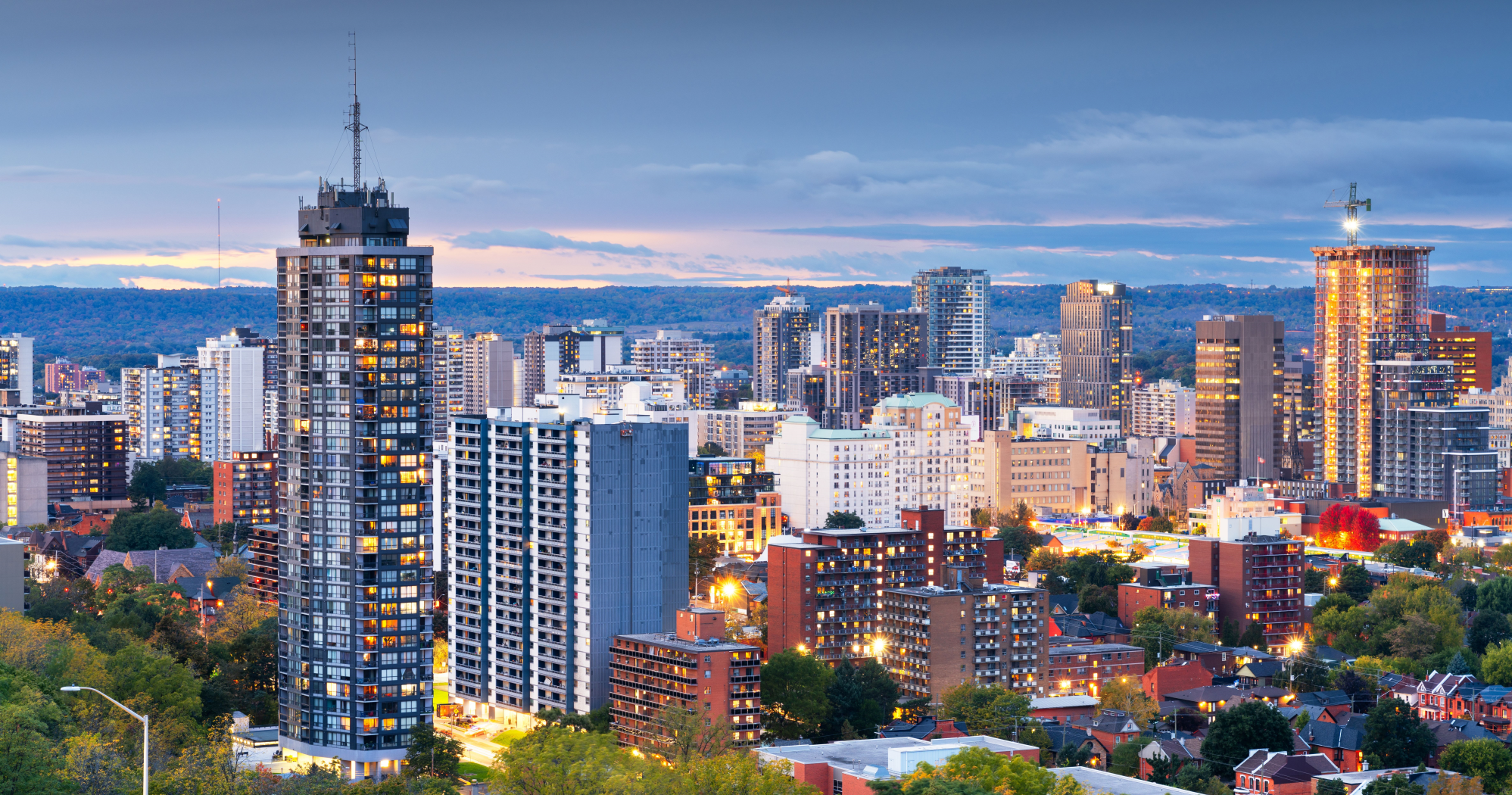 Downtown Hamilton cityscape at dusk featuring high-rise buildings, illuminated windows, and green trees in the foreground under a cloudy sky.