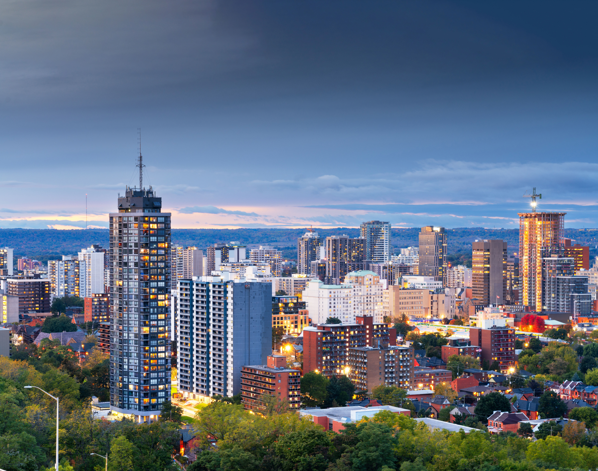 Cityscape at dusk featuring high-rise buildings, illuminated windows, and green trees in the foreground under a cloudy sky.
