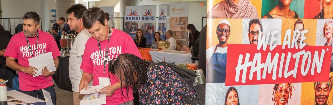 People visit informational booths at a community event; a “We Are Hamilton” sign is displayed in the foreground.