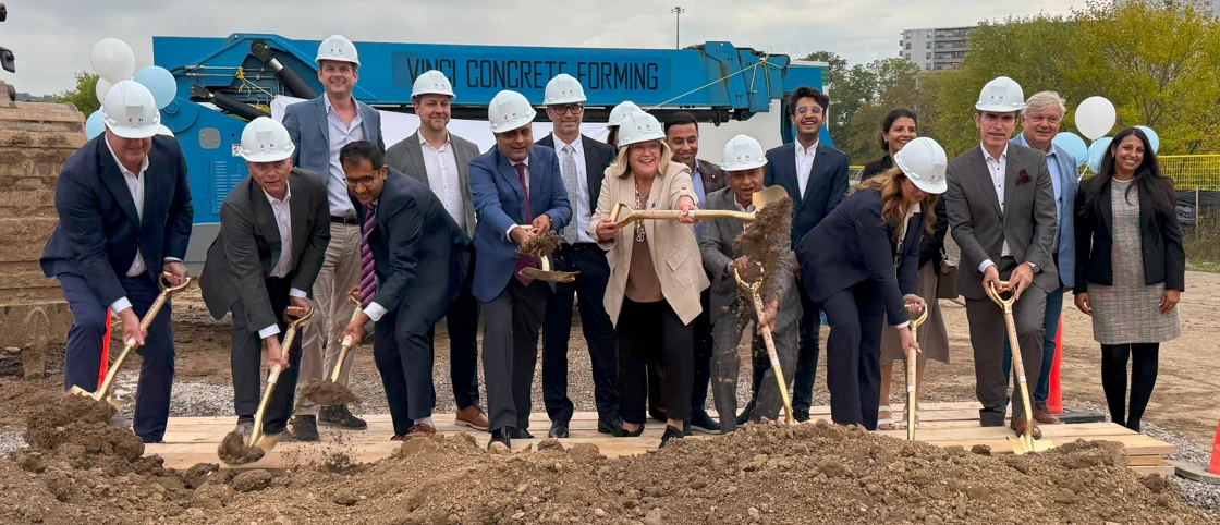 People in business attire shoveling dirt during a ceremonial ribbon-cutting