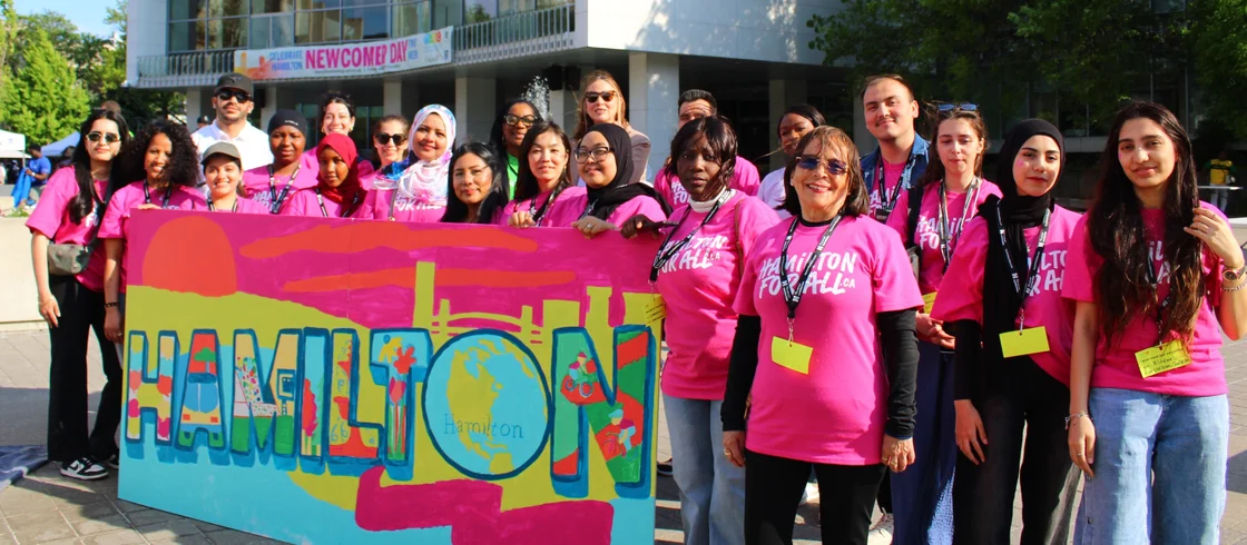 A group of volunteers and staff from Hamilton Immigration Partnership Council post with a Hamilton-themed mural painting in front of Hamilton City Hall during the annual Newcomer Day event
