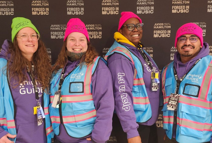 Four volunteer Night Guides dressed in toques and vests, standing in front of a Junos Hamilton: Forged in Music pull-up banner