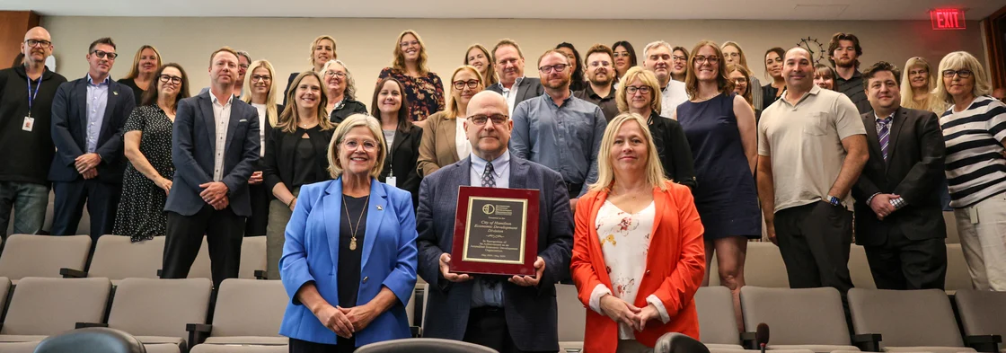 The City of Hamilton's Economic Development Division staff posing for a picture in the city council chambers while holding up a plaque highlighting that the division is an Accredited Economic Development Organization.