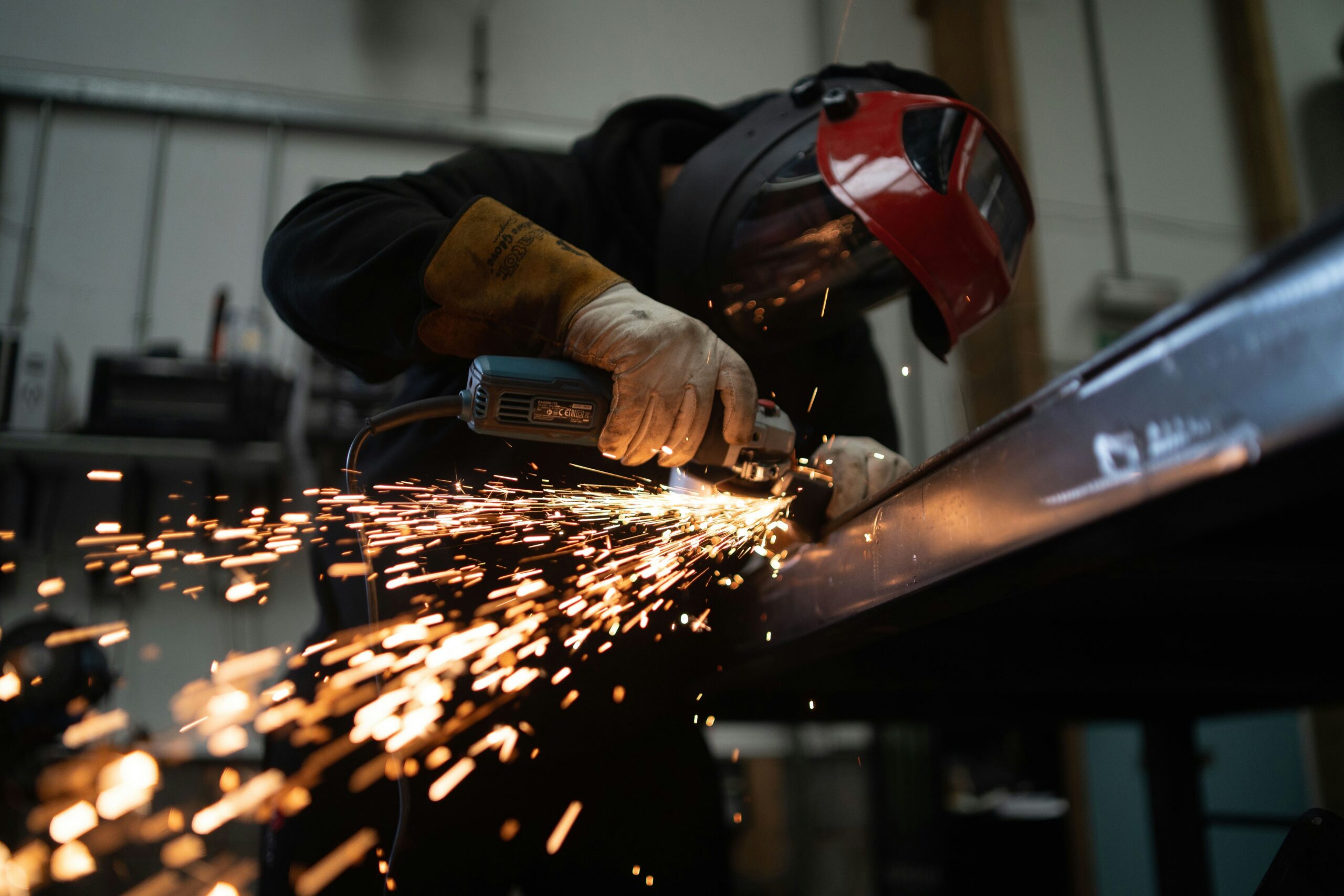A welder using a welding tool inside of an industrial building. They're wearing welding equipment and bright sparks can be seen.