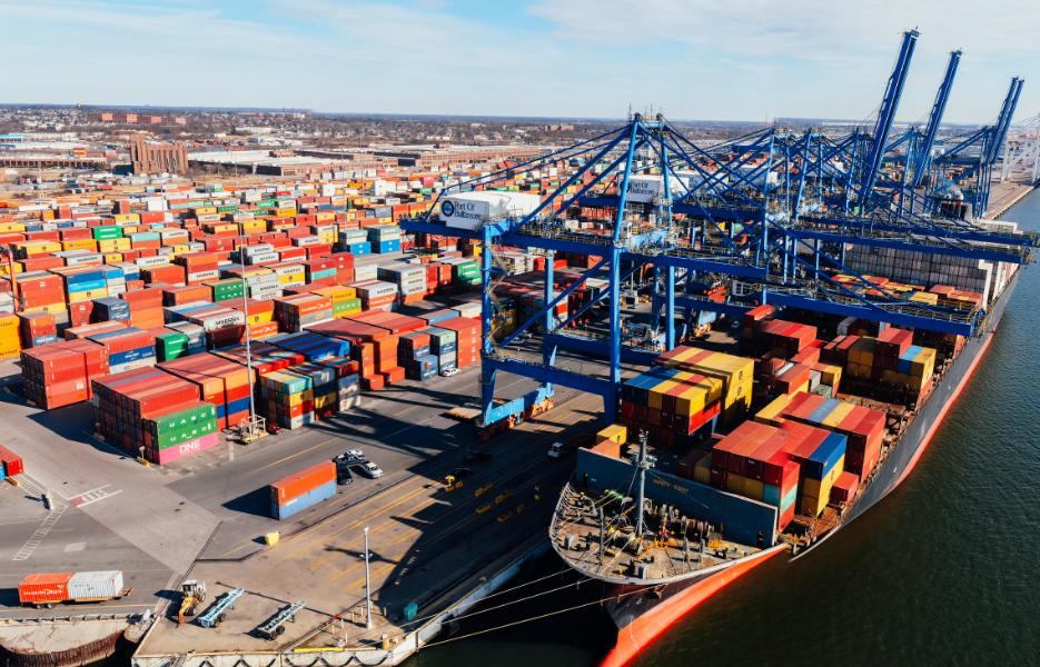 Industrial land, covered in multi-coloured shipping containers. Containers can be seen being loaded onto a large cargo ship docked at a port.