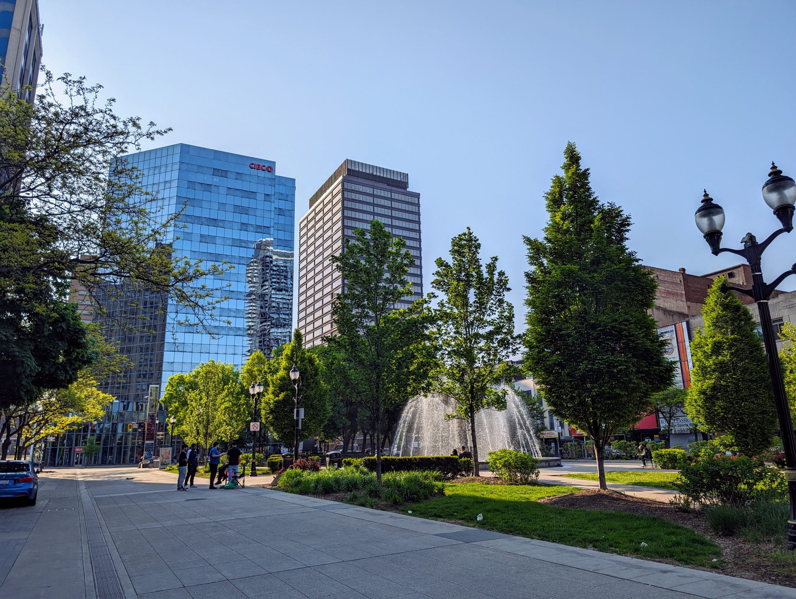 A wide, west-facing summer shot of Gore Park in Hamilton. The fountain is activated, and people can be seen standing off to the side, surrounded by trees. Tall buildings can be seen in the background.