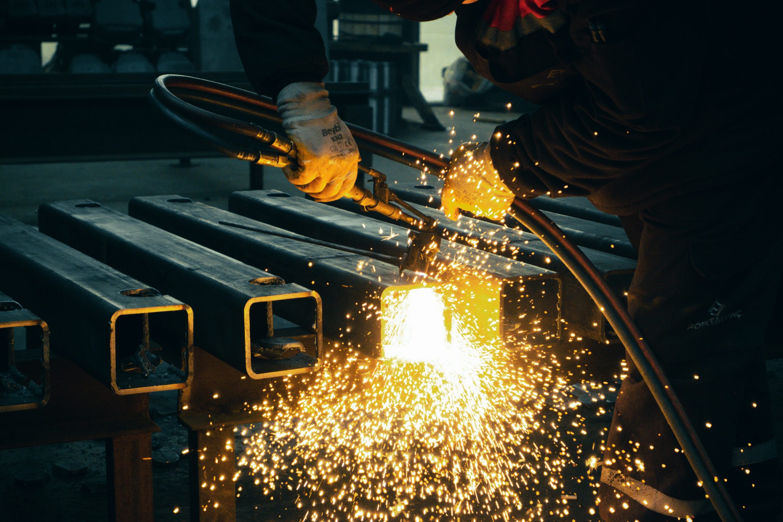 A steel worker working in a dark factory floor with protective gear and sparks flying from his tool